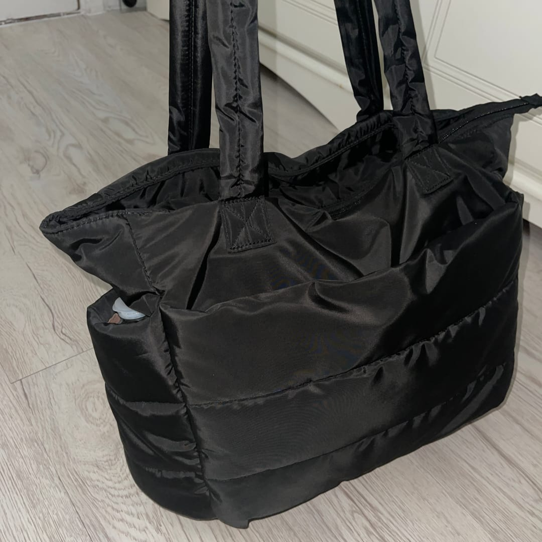 „Czarna pikowana torba stojąca na jasnej podłodze, widoczny bok i wnętrze torby; elegancki połysk materiału.”