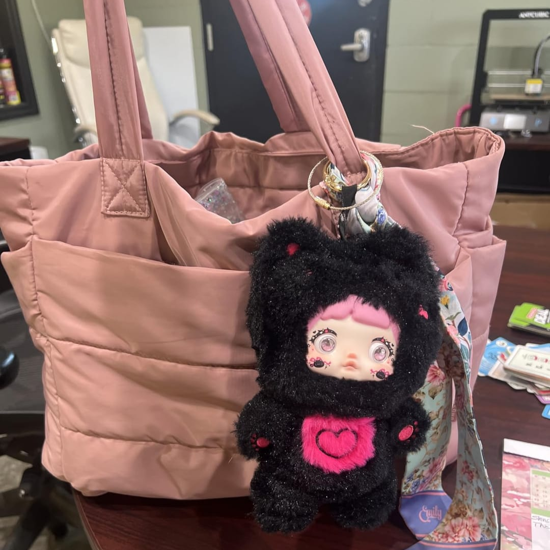 „Różowa pikowana torba stojąca na stole, ozdobiona czarną maskotką z różowym sercem i kolorowym brelokiem; wnętrze torby częściowo otwarte.”