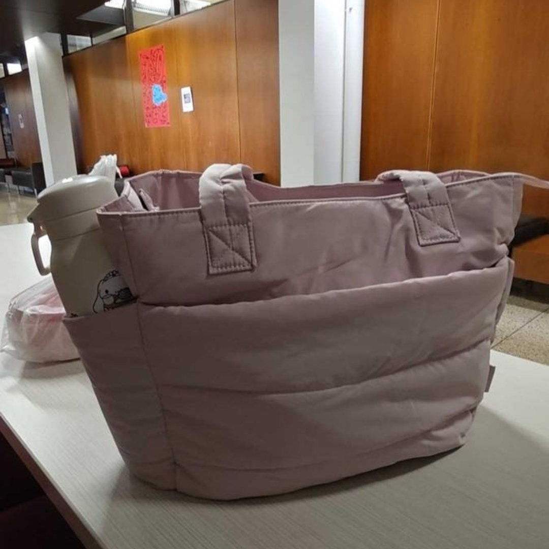 „Różowa pikowana torba termiczna stojąca na stole, z białym kubkiem w bocznej kieszeni; widoczne wnętrze pomieszczenia w tle.”