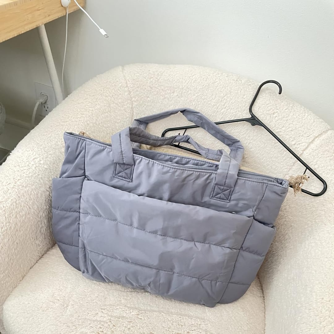 „Szara pikowana torba położona na kremowym fotelu, obok czarnego wieszaka; jasne, minimalistyczne wnętrze w tle.”