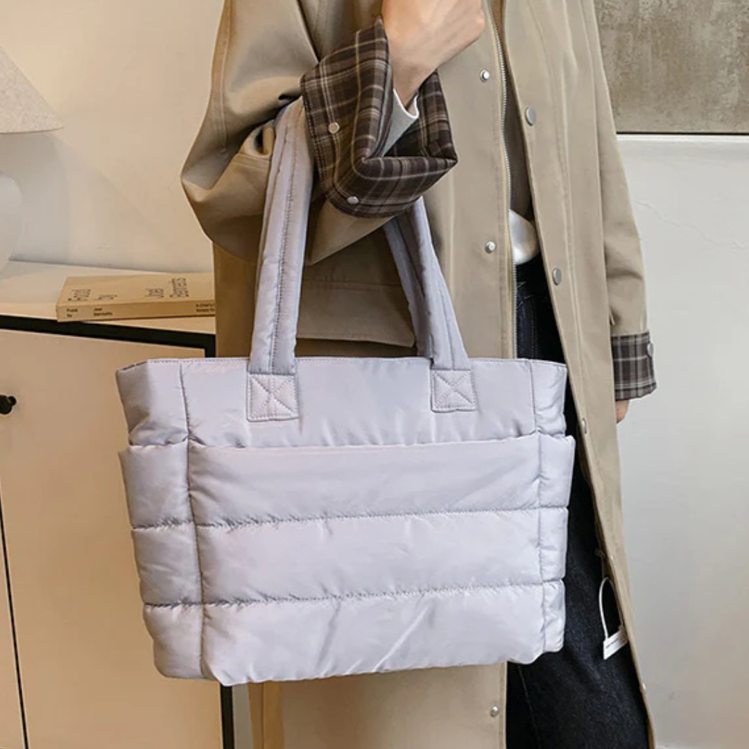 „Szara pikowana torba na ramię trzymana przez osobę w beżowym płaszczu z podwiniętymi rękawami; widoczne wnętrze pokoju w tle.”