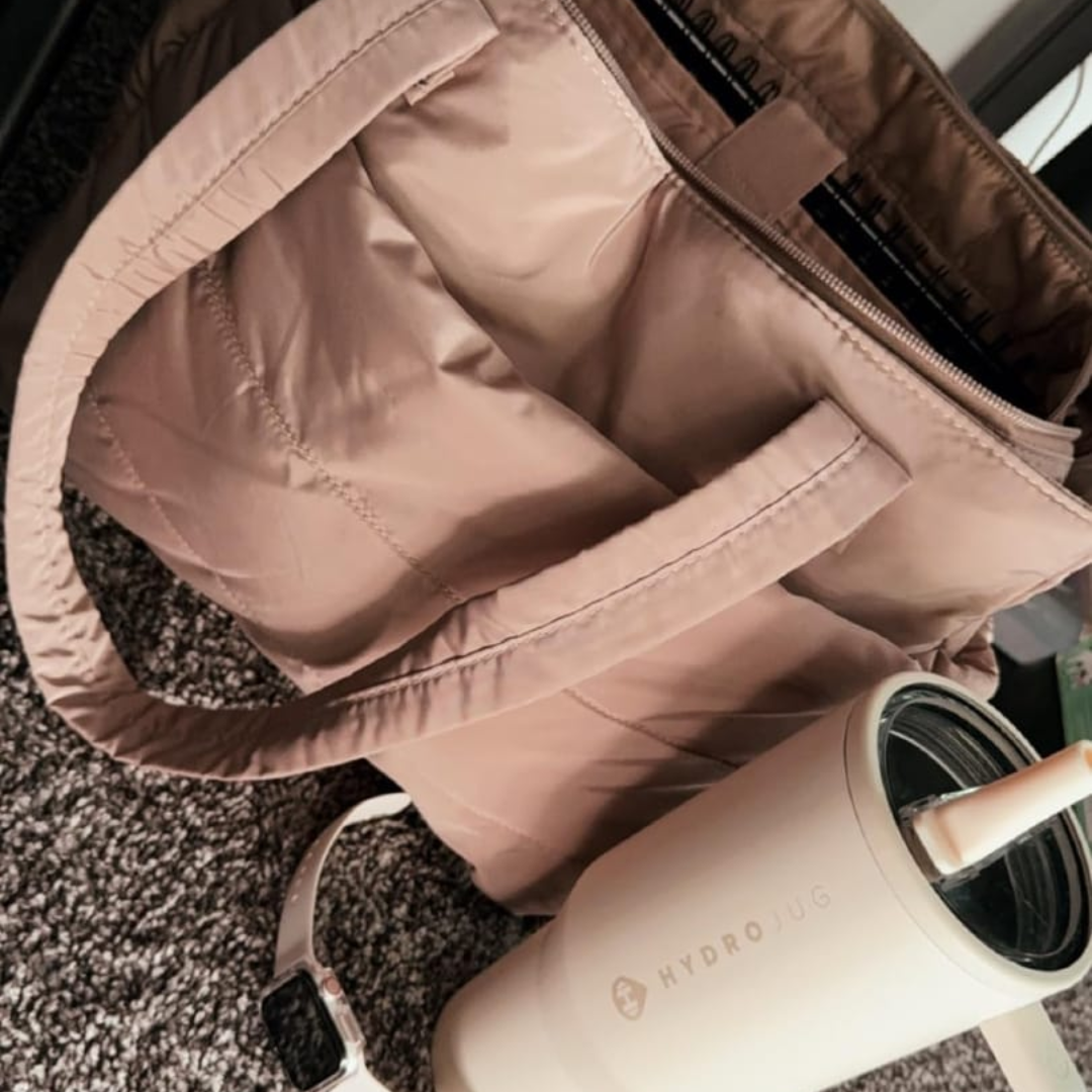 „Beżowa torba Softi na dywanie, otwarta z widocznym notesem; obok jasny kubek/bidon ze słomką i zegarek.”