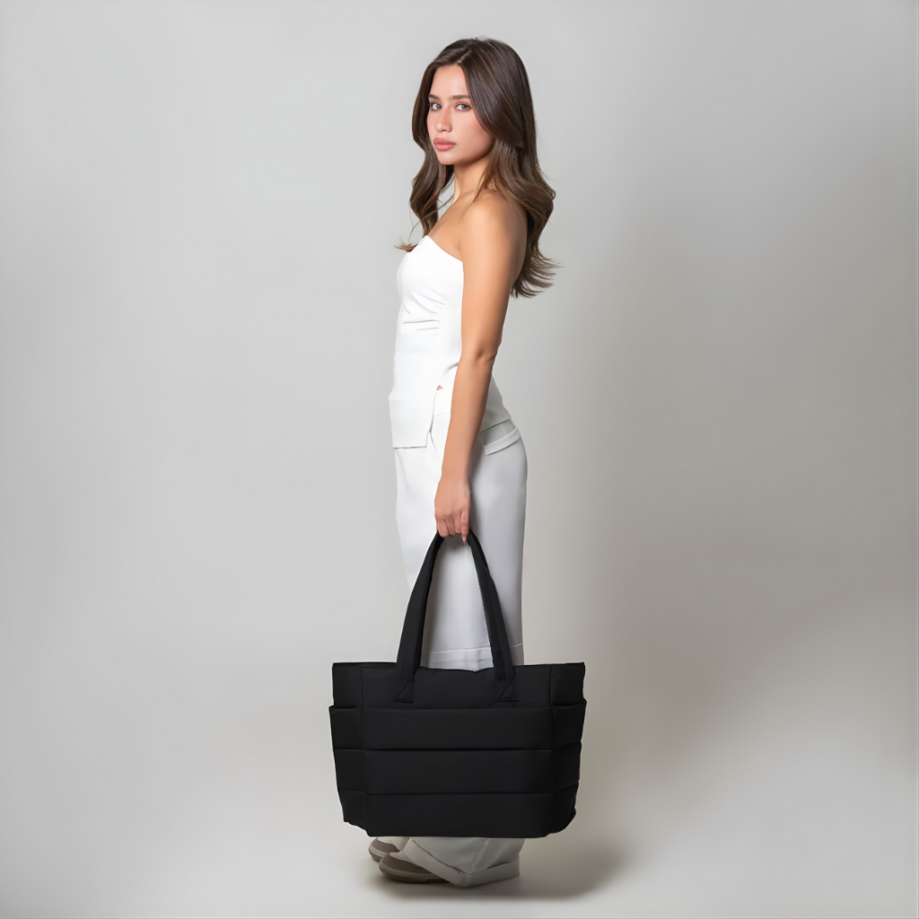 „Modelka w białym stroju trzyma czarną torbę Softi w dłoni, widok z boku na neutralnym, jasnym tle; duża, pikowana torba z długimi uchwytami.”