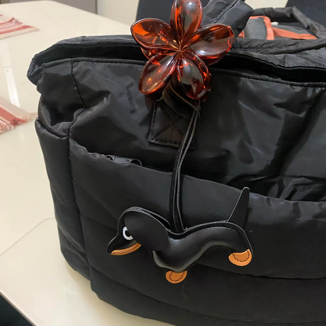 „Czarna torba Softi w pomieszczeniu; na przedniej kieszeni zawieszki – kwiat i czarna kaczka z pomaranczowymi detalami.”