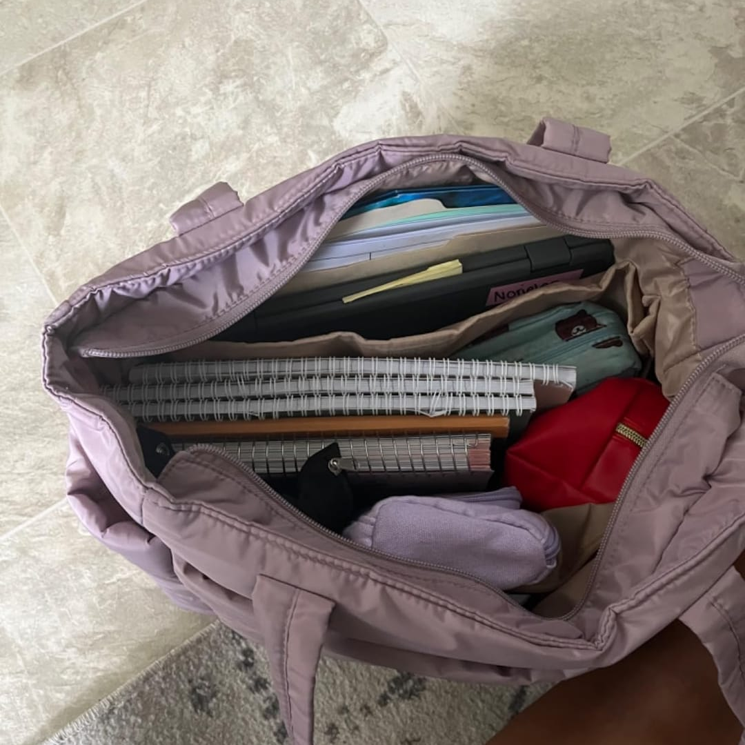 „Lawendowa torba Softi otwarta z góry; w środku spiralne zeszyty, laptop, czerwone etui i ubranie, widoczne przegrody organizera.”