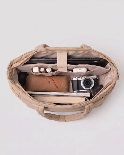 Widok z góry na otwarte wnętrze beżowej torebki Softi. Ukazuje zorganizowaną przestrzeń z laptopem, aparatem fotograficznym, etui na okulary, książką, słuchawkami i drobnymi kosmetykami umieszczonymi w dedykowanych przegrodach.